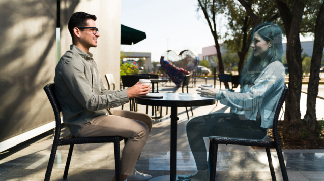 Happy young man with VR glasses and sitting at an outdoors coffee shop while talking with a virtual friend during a 3D simulation